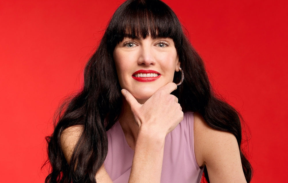Woman with long hair and red lipstick wearing a sleeveless top poses against a vibrant red background, showcasing elegant hoop earrings.