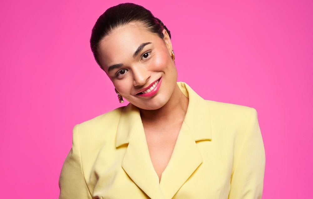 Woman in a yellow blazer with sleek hair, wearing gold earrings, smiling against a vibrant pink background.