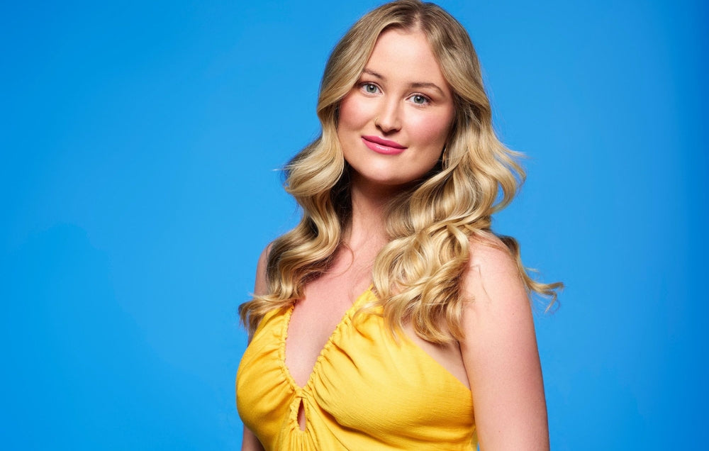 Smiling woman with long blonde hair in a yellow dress against a bright blue background.