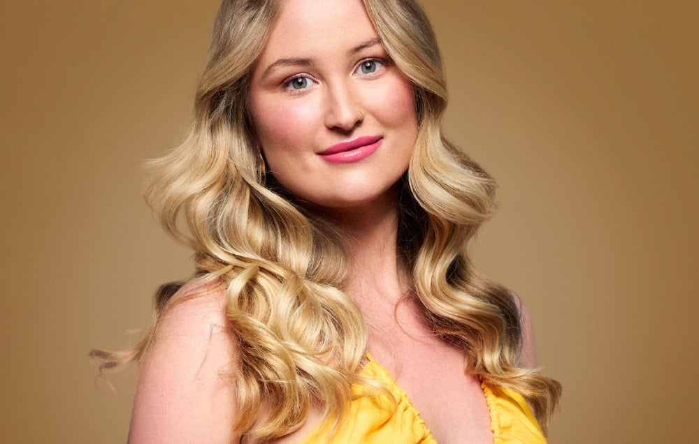 Woman with wavy blonde hair and pink lips, wearing a yellow top, against a warm beige background. Luxury jewellery theme.
