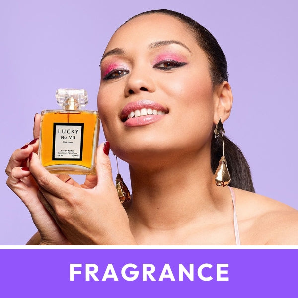 Woman smiling, holding a "Lucky No VII" perfume bottle against a light purple background. She wears statement earrings. Text: "FRAGRANCE".