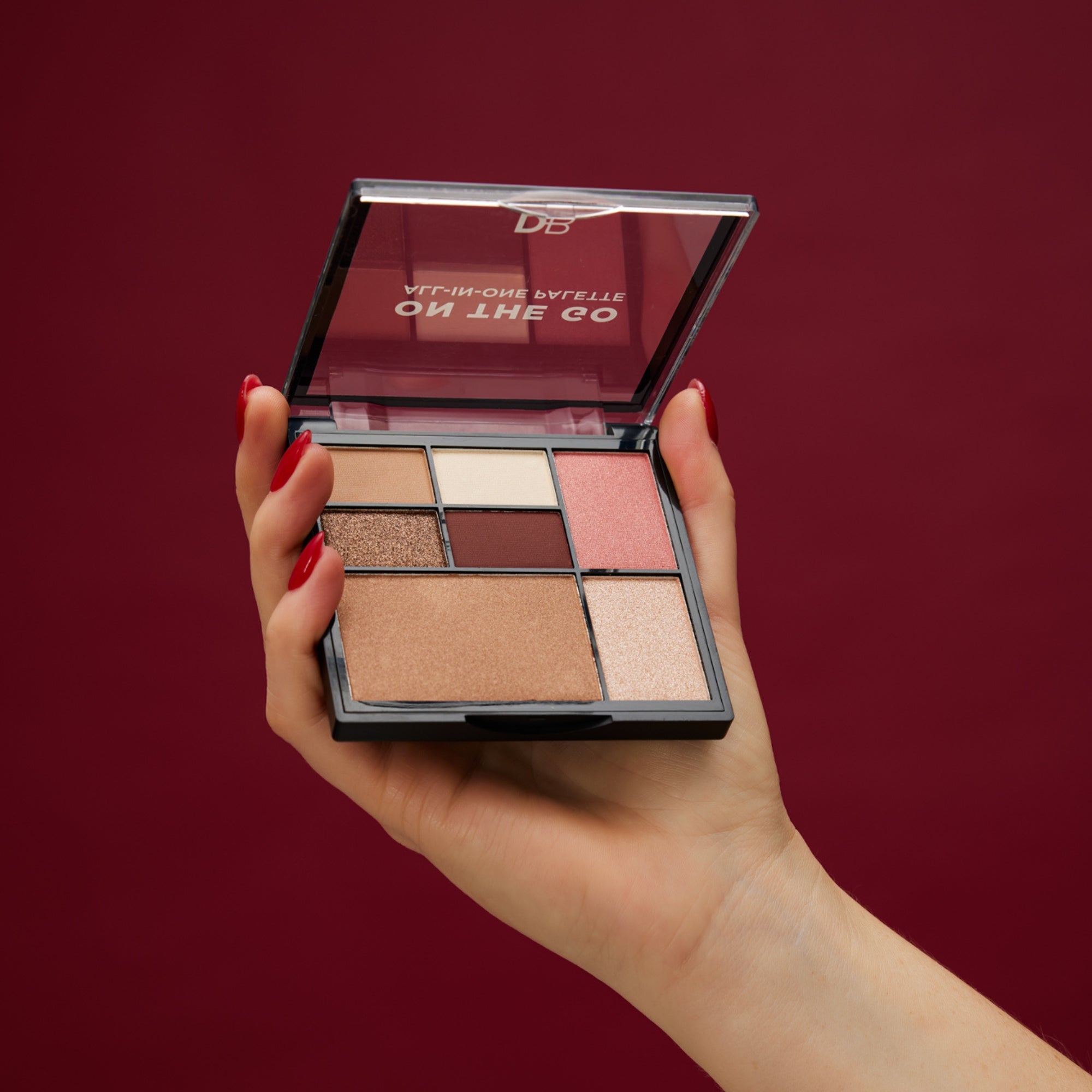 A hand holds an open makeup palette with nine shades of brown and pink, set against a deep burgundy background. Perfect for luxury beauty.
