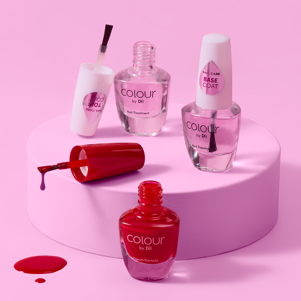 3x Colour by DB nail treatment bottles arranged on a pink cylindrical pedestal against a pink background. The set includes a nail polish bottle with brush, a base coat bottle, top coat & nail polish bottles, all in clear glass with white caps and pink labels. | colour-china-red