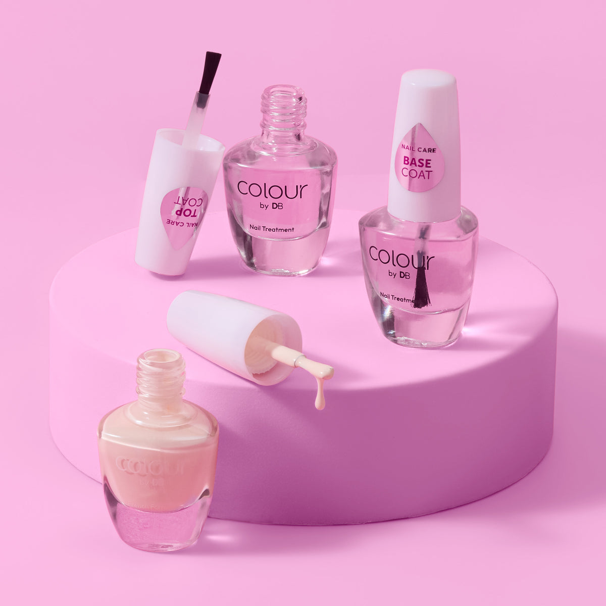 3x Colour by DB nail treatment bottles arranged on a pink cylindrical pedestal against a pink background. The set includes a nail polish bottle with brush, a base coat bottle, top coat & nail polish bottles, all in clear glass with white caps and pink labels. | colour-coconut-cream