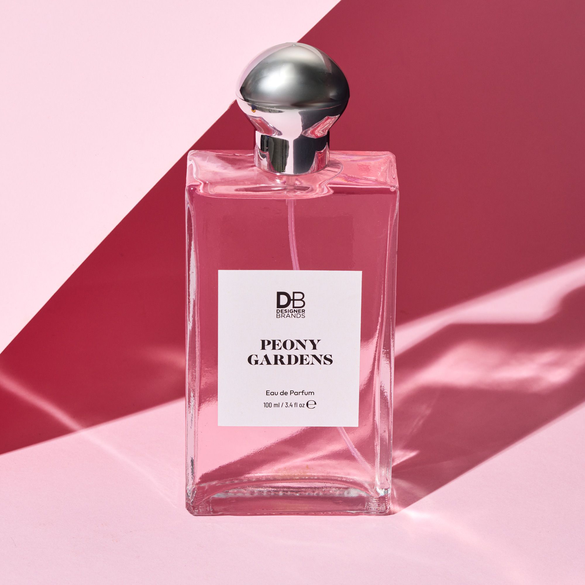 Elegant pink perfume bottle with silver cap, labelled "Peony Gardens" by DB Brands, against a two-tone pink background. Luxurious fragrance display.