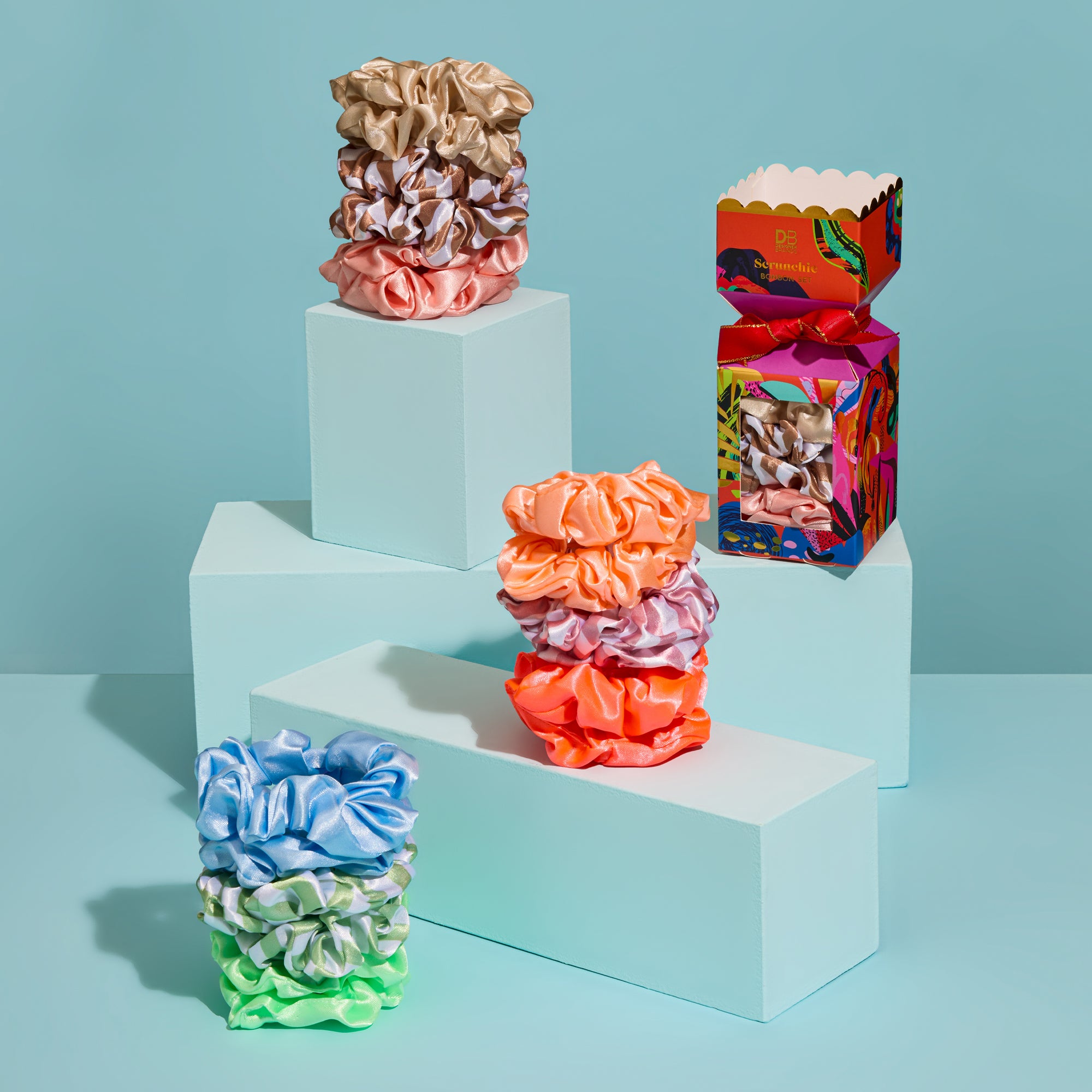 Colorful scrunchies on a geometric step structure with a colorful box against a light blue background