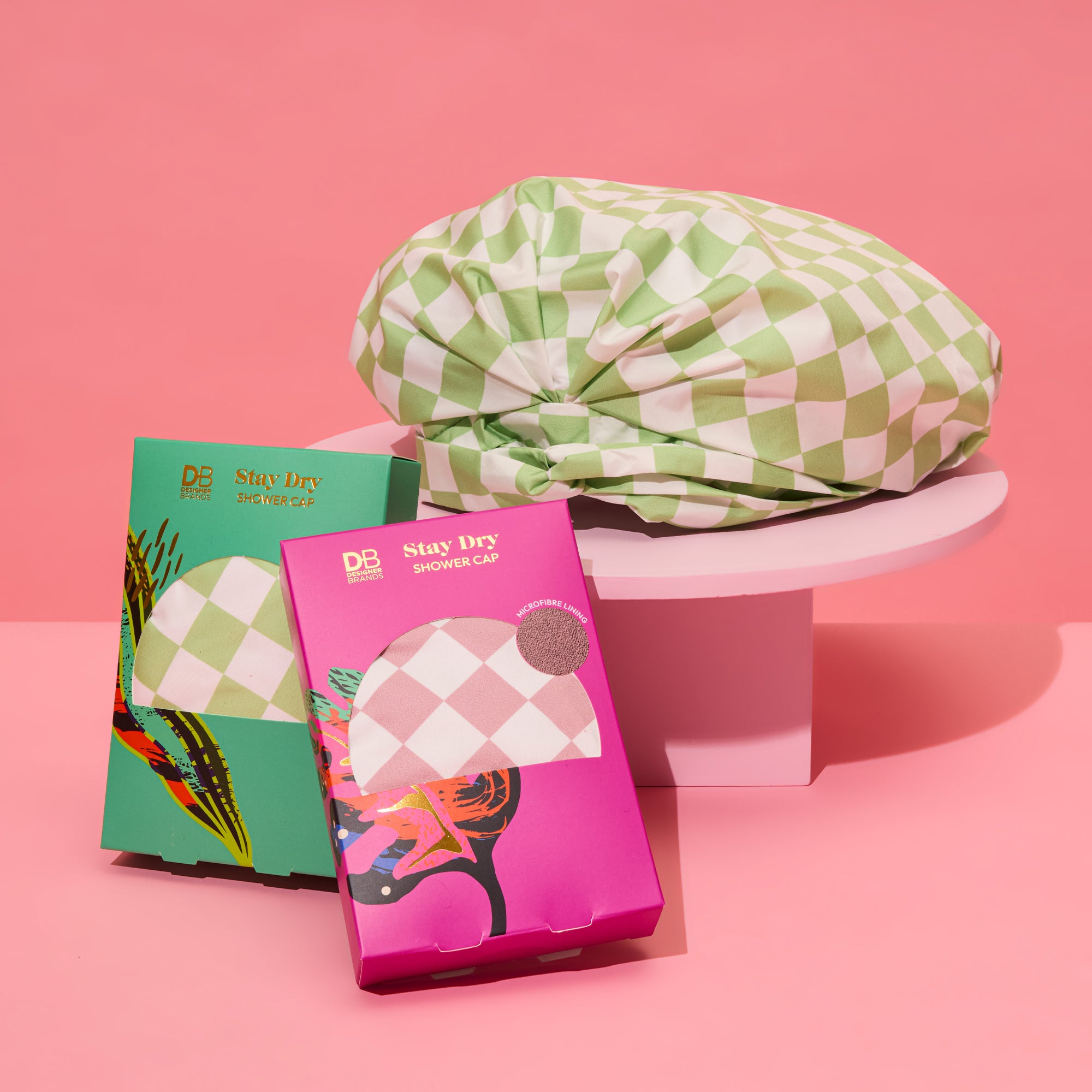 Green and white checkered shower cap on a pedestal, flanked by two decorative boxes in pink and teal, against a pink background.