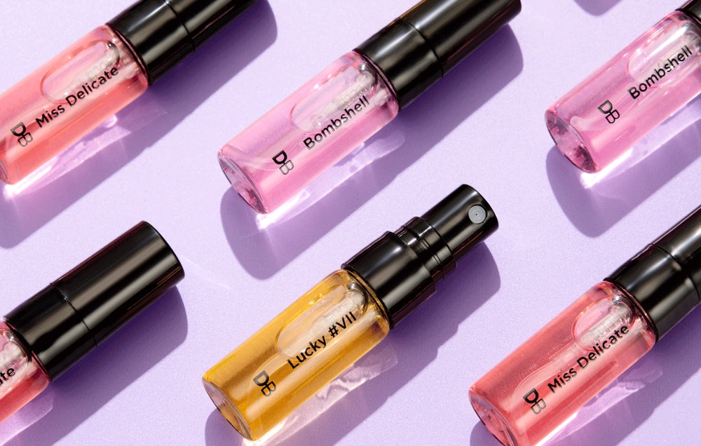 Elegant perfume vials arranged on a purple surface, featuring names like "Miss Delicate," "Bombshell," and "Lucky #VII" in luxurious packaging.