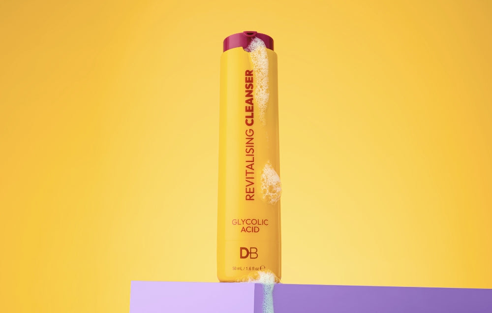 Revitalising Cleanser bottle with glycolic acid on a purple platform, set against a vibrant yellow background.